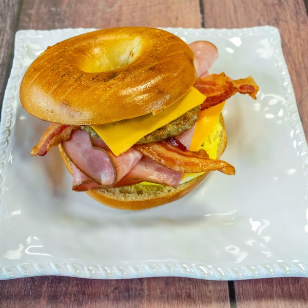 A Chelsea Coffee Company breakfast bagel with egg, cheese, sausage, ham, and bacon on a white plate.