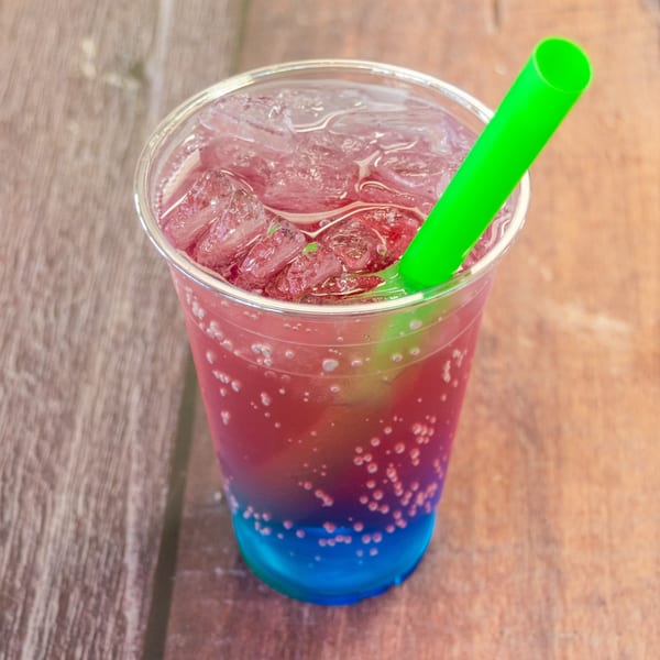 A vibrant, layered drink with red and blue liquid, ice, and a green straw on a wooden surface.