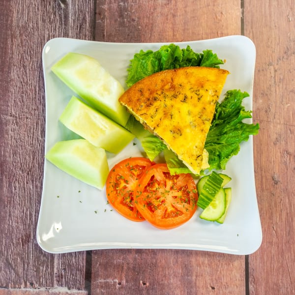 A slice of Chelsea Coffee Company's quiche with a side of honeydew melon, tomatoes, and cucumbers.