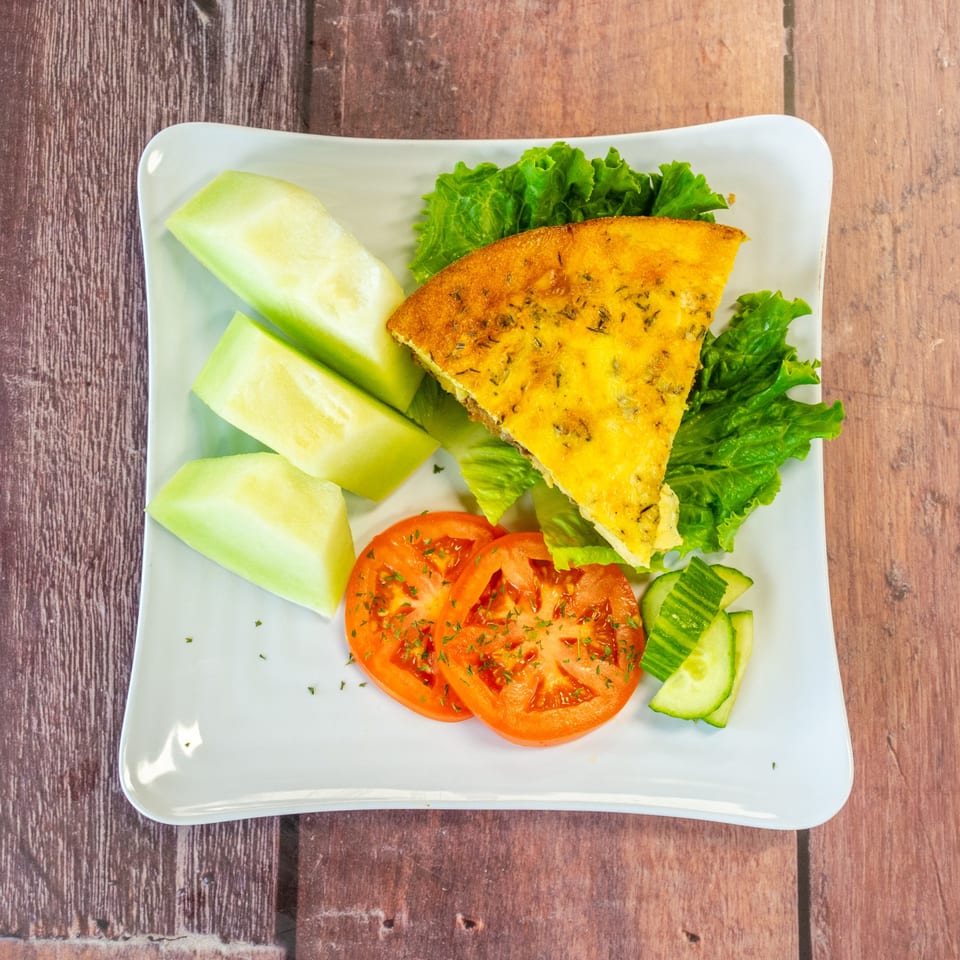 A slice of Chelsea Coffee Company's quiche with a side of honeydew melon, tomatoes, and cucumbers.